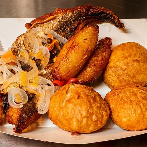 Fried fish, plantains, and dumplings with pickled onions in a white paper tray.