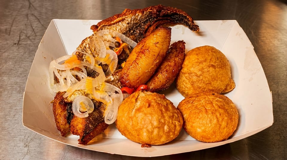Fried fish, plantains, and dumplings with pickled onions in a white paper tray.